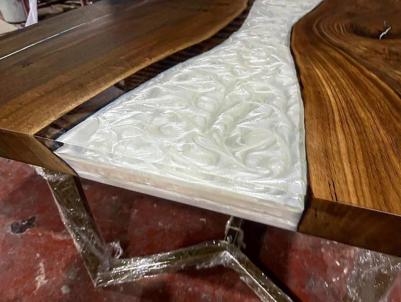 Puede incluir: Primer plano de una mesa de madera con un r&iacute;o de resina epoxi blanca que fluye por el centro. La resina tiene un patr&oacute;n de remolino y m&aacute;rmol. La mesa est&aacute; soportada por una base de metal.