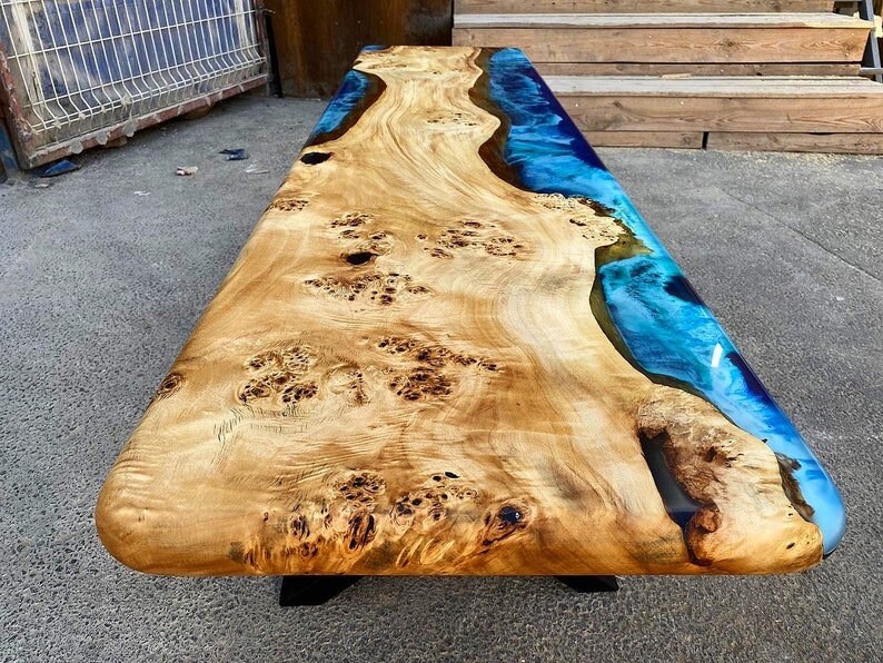 May include: A large wooden table with a blue epoxy resin inlay. The wood has a natural burl pattern and the resin is a deep blue color.