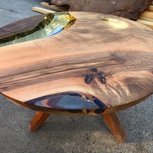 May include: Round wooden table with a live edge and a clear epoxy river running through the center. The table has three legs.