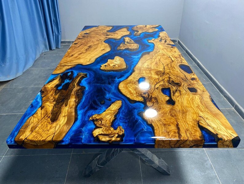 May include: A large wooden table top with a blue epoxy resin inlay. The resin has a flowing, river-like pattern. The wood is a light brown color with dark brown streaks.