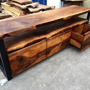 May include: A wooden entertainment center with a live edge top and three drawers. The drawers are made of wood with a dark brown finish. The entertainment center is made of black metal and wood.