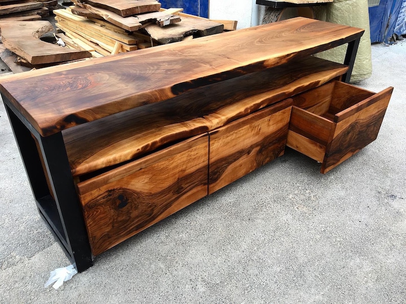 May include: A wooden entertainment center with a live edge top and three drawers. The drawers are made of wood with a dark brown finish. The entertainment center is made of black metal and wood.