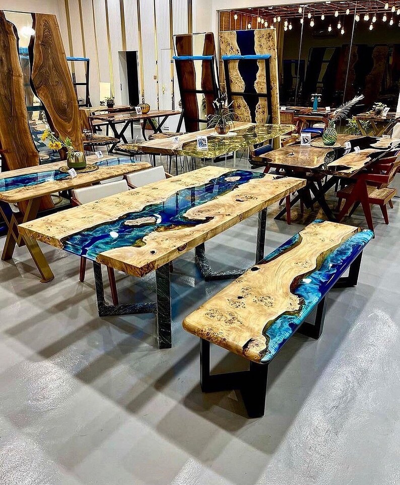 May include: A large wooden table with a blue and green epoxy resin river flowing through the center. The table has black metal legs and is paired with a matching bench. The table and bench are made of wood with a natural grain pattern.