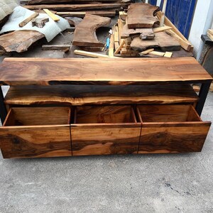 May include: A wooden entertainment center with a live edge top and three drawers. The drawers are made of wood and have a black metal frame.