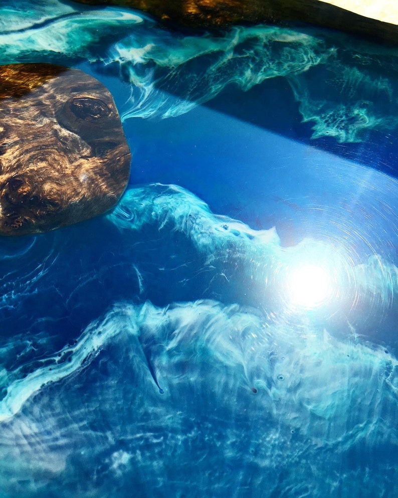 May include: A close-up of a blue and white resin table top with a dark wood inlay. The resin has a swirling pattern that resembles water.