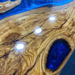 May include: Close-up of a wooden table top with a blue epoxy resin inlay. The wood has a natural grain pattern and the resin is smooth and shiny. The table top is illuminated by spotlights, creating a reflective surface.