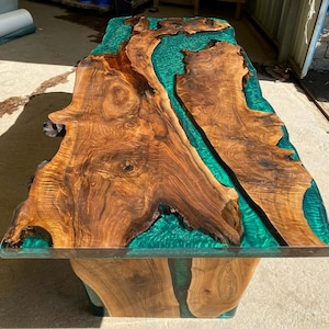 Könnte beinhalten: Handgefertigter Holztisch mit Live-Edge-Design. Der Tisch hat eine reichhaltige, braune Holzmaserung mit einem lebendigen türkisfarbenen Epoxidharzfluss. Der Tisch wird von einem passenden Holzsockel getragen, was ein einzigartiges Möbelstück ergibt.