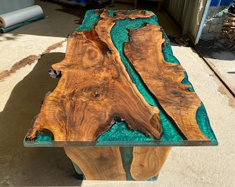 Può includere: Tavolo in legno fatto a mano con design a bordo vivo. Il tavolo presenta un ricco piano in legno marrone con un vibrante fiume in resina epossidica turchese. Il tavolo è sostenuto da una base in legno abbinata, creando un pezzo unico.