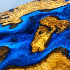 May include: A close-up of a wooden table top with a blue epoxy resin river running through it. The wood has a natural grain pattern and the resin is smooth and shiny.