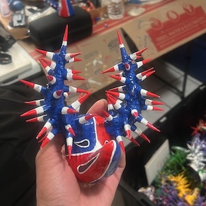May include: A handmade, colorful sculpture held in a person's hand. The sculpture is primarily blue with white and red accents, featuring a spiky design. The base is red and blue with a white design. Other similar sculptures are visible in the background.