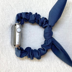 May include: A navy blue scrunchie watch band with a bow on the end. The band is made of soft, stretchy fabric and is designed to fit most Apple Watch sizes.