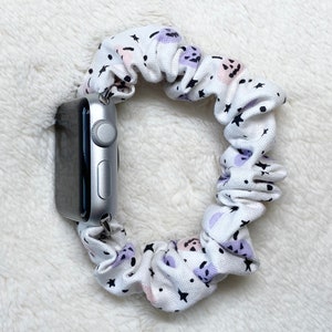 May include: White scrunchie watch band with a pattern of black stars, purple shapes, and pink dots. The band is attached to a silver smartwatch. The scrunchie is made of a soft fabric.