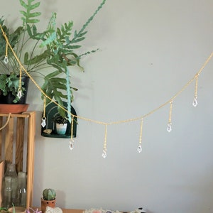 May include: A gold beaded garland with clear crystal teardrop pendants hangs against a white wall. The garland is decorated with small, clear crystals. The garland is hanging in front of a green plant.