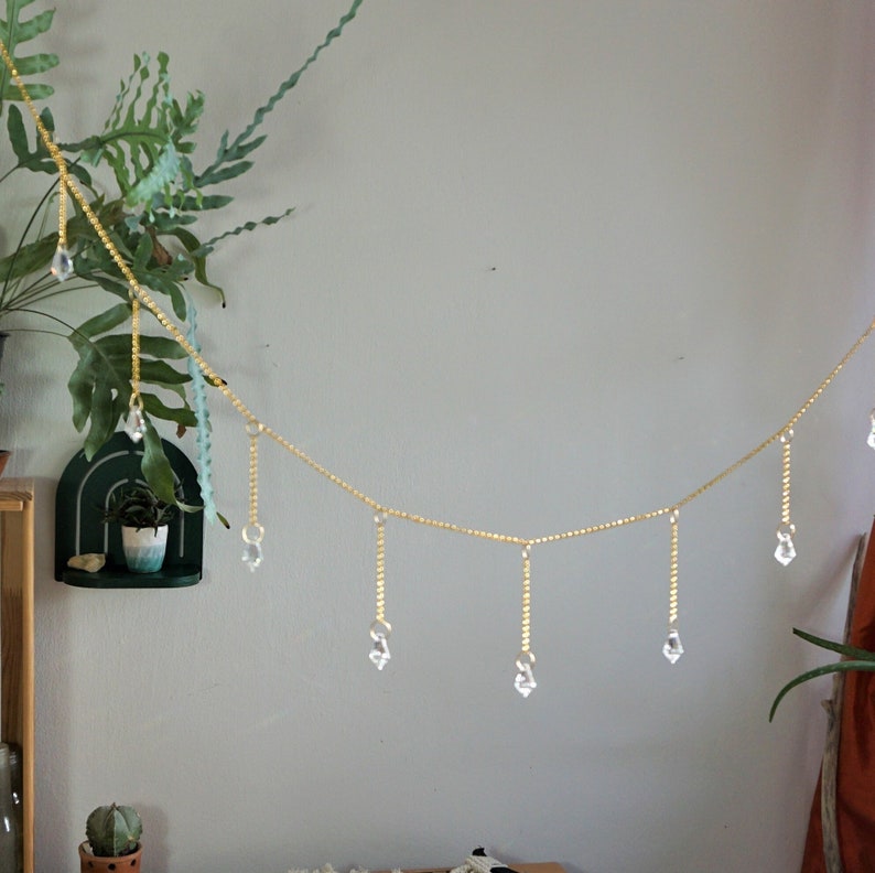 May include: A gold beaded garland with crystal drops hanging from it. The garland is strung across a white wall.