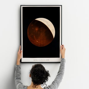 May include: A framed black and white photograph of a partial eclipse of the moon. The moon is a reddish-brown color with a sliver of white light on the right side. The text "PARTIAL ECLIPSE of the MOON" is printed below the image.