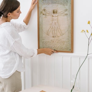 Może przedstawiać: Kobieta wiesza na białej ścianie oprawiony wydruk &bdquo;Człowieka witruwiańskiego&rdquo; Leonarda da Vinci. Wydruk znajduje się w drewnianej ramie i przedstawia szczeg&oacute;łowy rysunek nagiej postaci męskiej w okręgu i kwadracie.