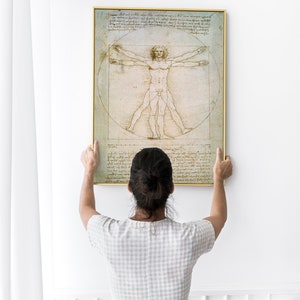 Może przedstawiać: Osoba wiesza na białej ścianie oprawiony wydruk &bdquo;Człowieka witruwiańskiego&rdquo; Leonarda da Vinci. Wydruk jest czarno-biały i przedstawia szczeg&oacute;łowy rysunek nagiej postaci męskiej w okręgu i kwadracie.