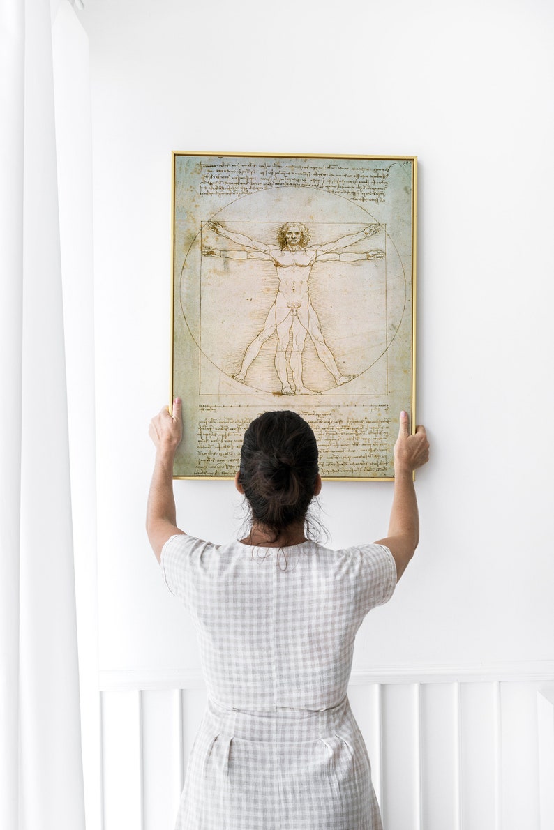 Może przedstawiać: Osoba wiesza na białej ścianie oprawiony wydruk &bdquo;Człowieka witruwiańskiego&rdquo; Leonarda da Vinci. Wydruk jest czarno-biały i przedstawia szczeg&oacute;łowy rysunek nagiej postaci męskiej w okręgu i kwadracie.