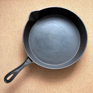May include: A black cast iron skillet with a long handle and a loop at the end. The skillet has a round cooking surface and a small pouring spout. The pan is sitting on a light brown surface.