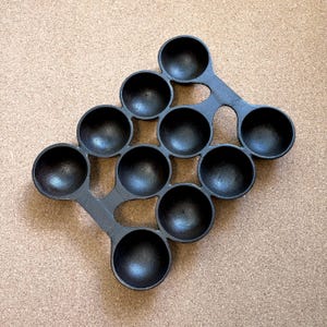 May include: A black cast iron pan with 10 circular indentations, connected by curved supports. The pan is designed for cooking multiple small, round items. The pan sits on a light brown surface.