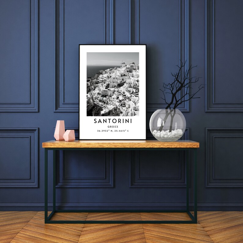 Puede incluir: Fotograf&iacute;a en blanco y negro de Santorini, Grecia. La imagen muestra una ciudad con edificios blancos y un oc&eacute;ano azul en el fondo. El texto "SANTORINI" est&aacute; impreso debajo de la imagen, junto con las coordenadas "36.3932&deg; N, 25.4615&deg; E".