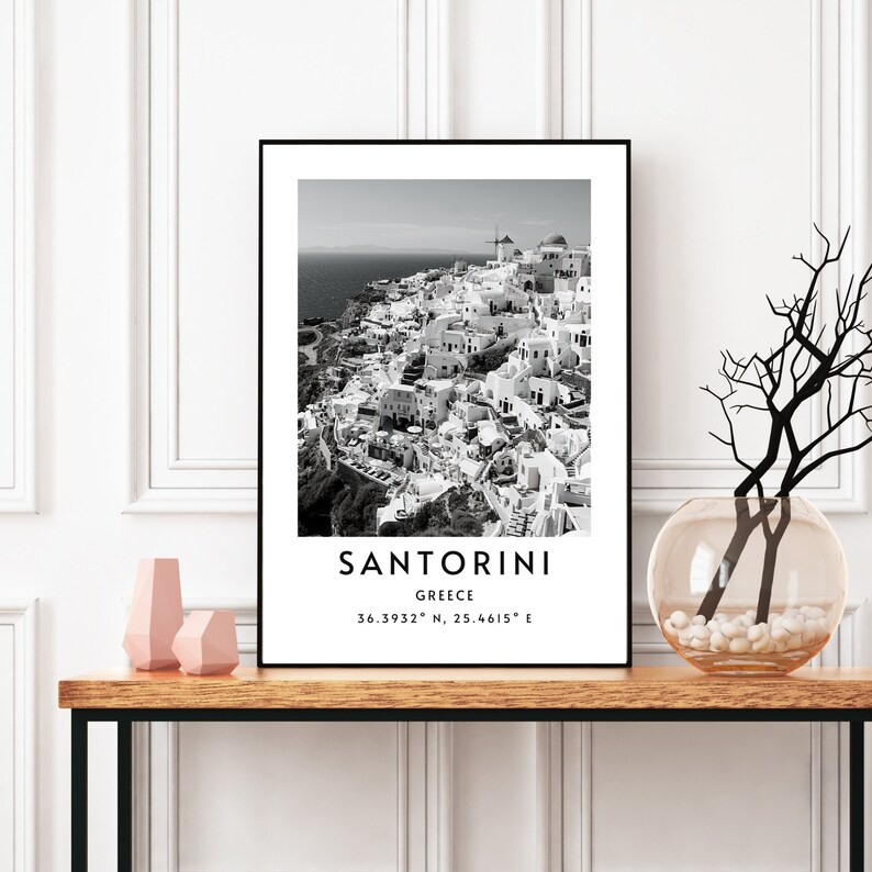 Puede incluir: Fotograf&iacute;a en blanco y negro de Santorini, Grecia. La imagen muestra una ciudad con edificios blancos agrupados en una ladera. El texto "Santorini, Grecia 36.3932&deg; N, 25.4615&deg; E" est&aacute; impreso debajo de la imagen.