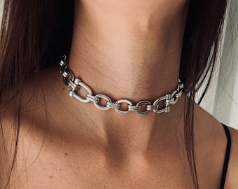 May include: A silver-toned choker necklace featuring a series of interlocking oval links connected by smaller, rectangular-shaped metal bars. The chain sits snugly around the neck, showcasing a polished metallic finish and a repeating geometric link pattern.