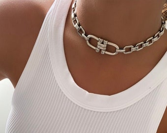 May include: A white ribbed tank top with a thick crew neckline paired with a chunky silver-toned metal chain link necklace and light blue denim jeans. The tank top features a fitted silhouette and vertical ribbed texture throughout the fabric.