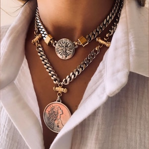 May include: A layered necklace set featuring two silver-toned curb chains. The shorter chain has a round pendant with a dove and the text SANCTE SPIRITUS. The longer chain features a round pendant depicting a profile of a person and the word JUSTICE.