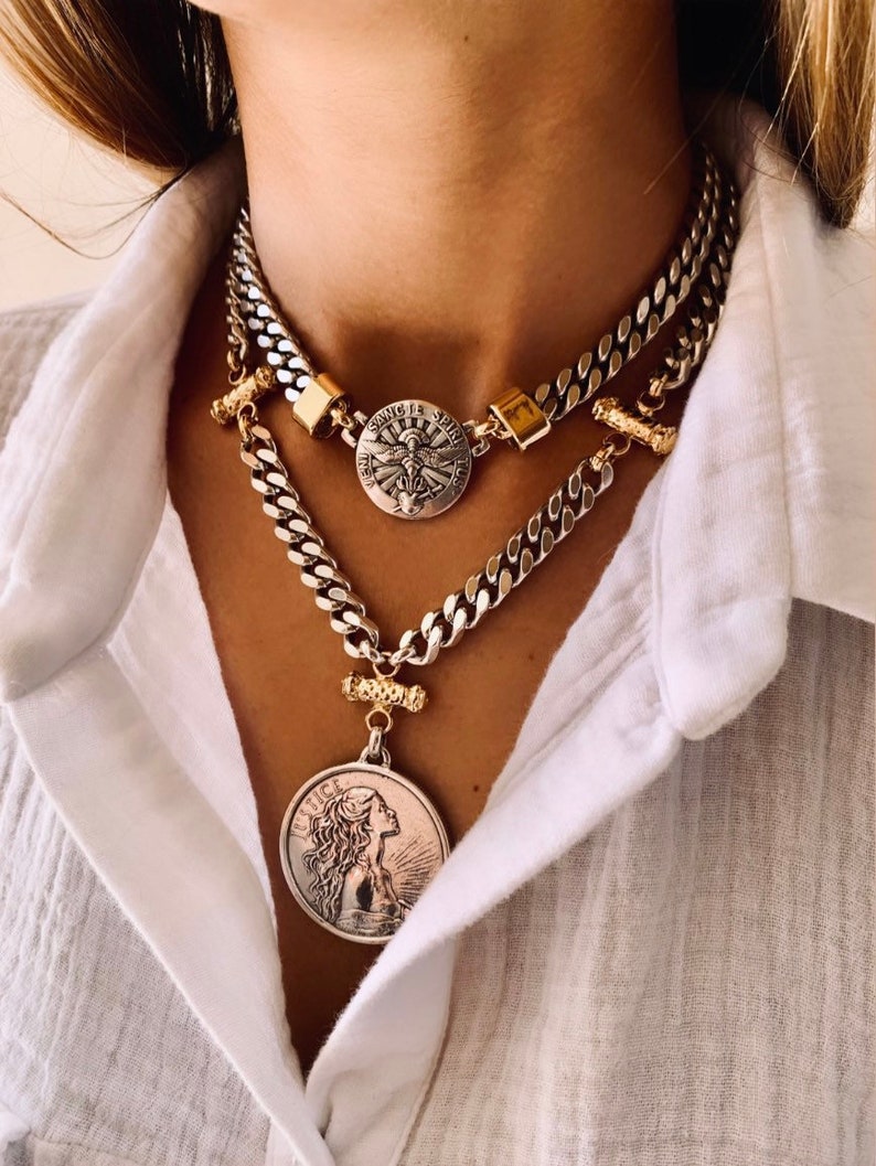 May include: A layered silver-tone chain necklace featuring two circular pendants. The top pendant displays a winged figure with the text Veni Sancte Spiritus. The lower, larger pendant depicts a profile of a figure with the word Justice inscribed.