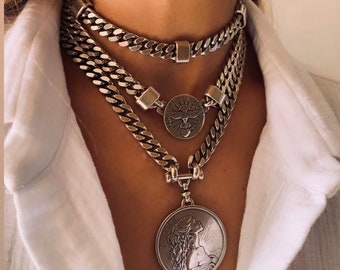 May include: A layered silver-toned chain necklace set featuring three distinct strands. The top strand is a plain curb chain, the middle features a circular pendant with a bird emblem, and the bottom includes a large coin pendant with a profile portrait.