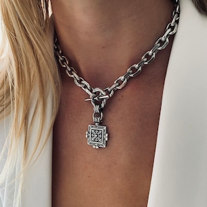 Op de afbeelding: Een zilveren ketting met een vierkante hanger met een ingewikkeld ontwerp. De hanger is met een zilveren ring aan de ketting bevestigd.