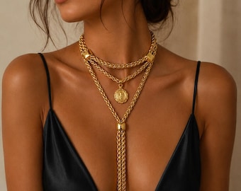 Puede incluir: Un collar de cadenas múltiples en tono dorado con un colgante circular de moneda y dos conectores metálicos cuadrados. El diseño incluye un elemento de cadena largo y colgante que se extiende hacia abajo desde una cuenta metálica central.