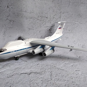 Op de afbeelding: Een wit en blauw modelvliegtuig met vier motoren en een hoge staartvin. Het model heeft de tekst "RA-76455" en "IL-76MD" erop gedrukt. De vleugels zijn grijs en het model staat op een grijze ondergrond.