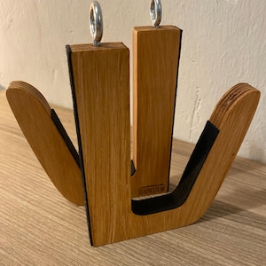 May include: Wooden surfboard rack with a natural wood grain finish. The rack has two vertical supports with metal rings at the top and curved arms with black padding. The rack is designed to hold surfboards.