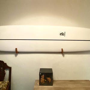 May include: A white surfboard with a black stripe down the center is mounted on a wall with wooden brackets. The surfboard has a logo near the center and is approximately 9 feet long.