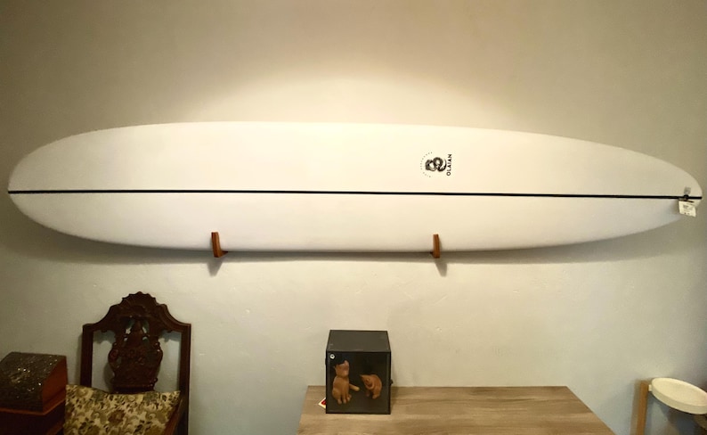 May include: A white surfboard with a black stripe down the center is mounted on a wall with wooden brackets. The surfboard has a logo near the center and is approximately 9 feet long.