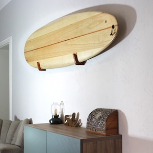 May include: A light-colored wooden surfboard mounted on a wall with two brown wooden brackets. The surfboard has a rounded shape and a thin, darker stripe down the center. A black loop is attached to the top of the board.