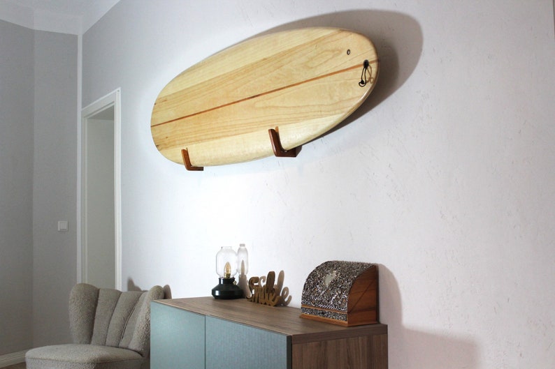 May include: A light-colored wooden surfboard mounted on a wall with two brown wooden brackets. The surfboard has a rounded shape and a thin, darker stripe down the center. A black loop is attached to the top of the board.