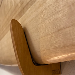 May include: A wooden surfboard rack with a curved design. The rack is made of light-colored wood and is mounted on a white wall. The surfboard is also made of light-colored wood. The rack has the text "BYNDSURF" on the bottom.