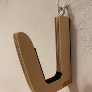 May include: A wooden surfboard wall rack with a curved design and black padding. The rack is mounted on a white wall with a metal hook and ring. The rack is made of light-colored wood.