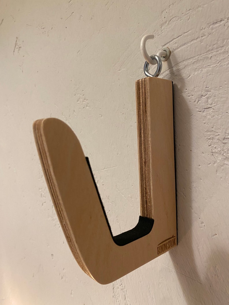 May include: A wooden surfboard wall rack with a curved design and black padding. The rack is mounted on a white wall with a metal hook and ring. The rack is made of light-colored wood.