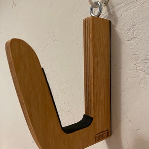May include: A wooden wall-mounted surfboard rack with a J-shaped design. The rack is made of light brown wood and features a black felt lining in the curve. A white hook and metal ring are used for hanging. The brand name "BRONGSPLNK" is visible.