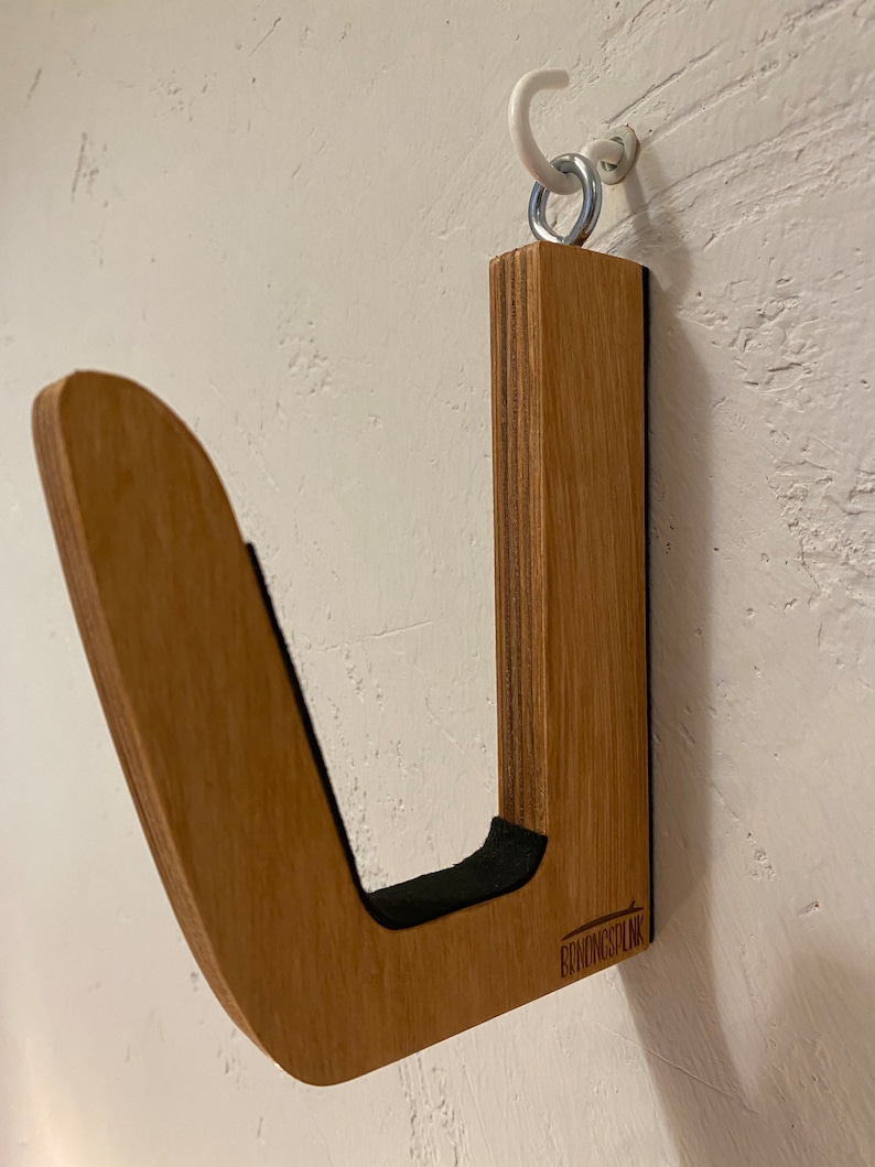 May include: A wooden wall-mounted surfboard rack with a J-shaped design. The rack is made of light brown wood and features a black felt lining in the curve. A white hook and metal ring are used for hanging. The brand name "BRONGSPLNK" is visible.