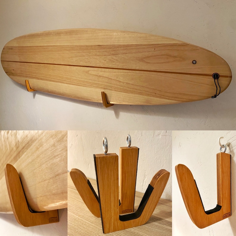 May include: A wooden surfboard displayed on a wall with two wooden surfboard racks. The surfboard is light brown with a dark brown stripe down the middle. The racks are made of wood and have a curved design.