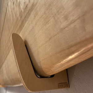 May include: A light-colored wooden surfboard mounted on a custom-made wooden wall rack. The rack is designed to hold the board securely against a wall. The rack has a curved design and is made of light-colored wood.