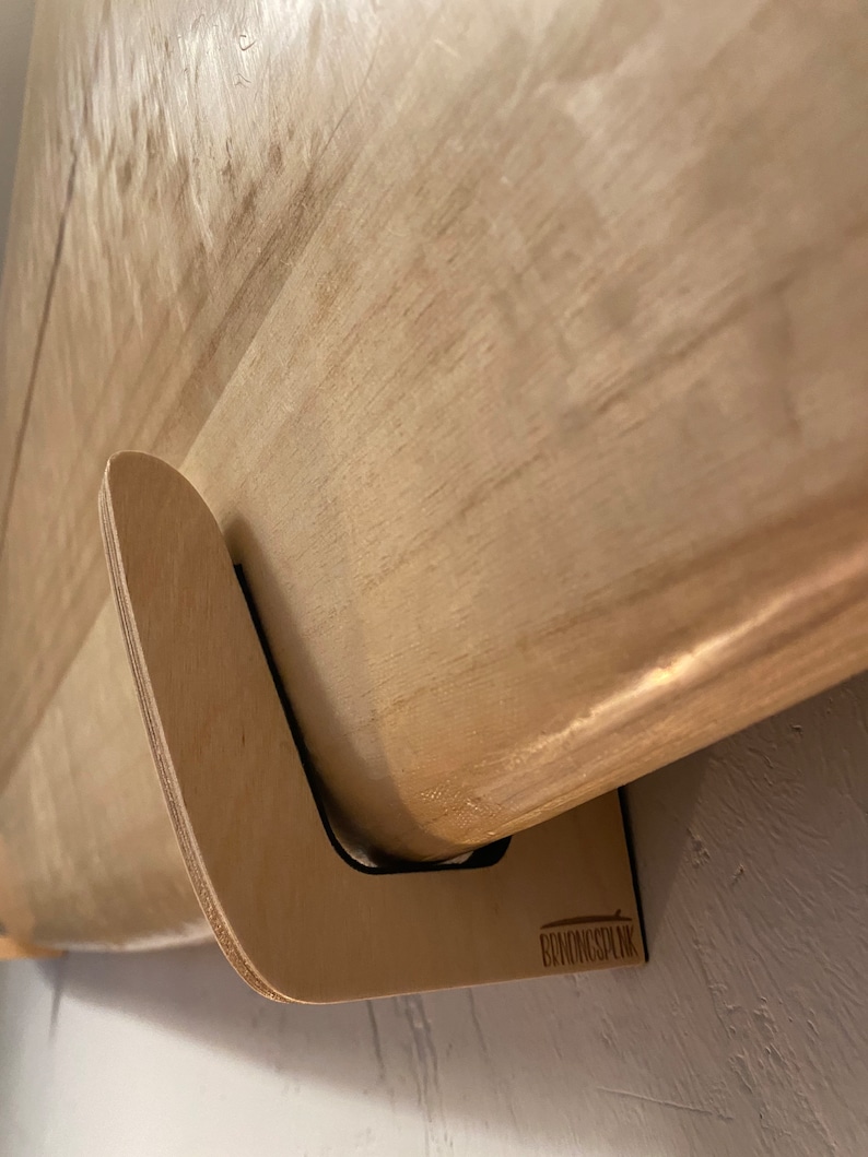 May include: A light-colored wooden surfboard mounted on a custom-made wooden wall rack. The rack is designed to hold the board securely against a wall. The rack has a curved design and is made of light-colored wood.