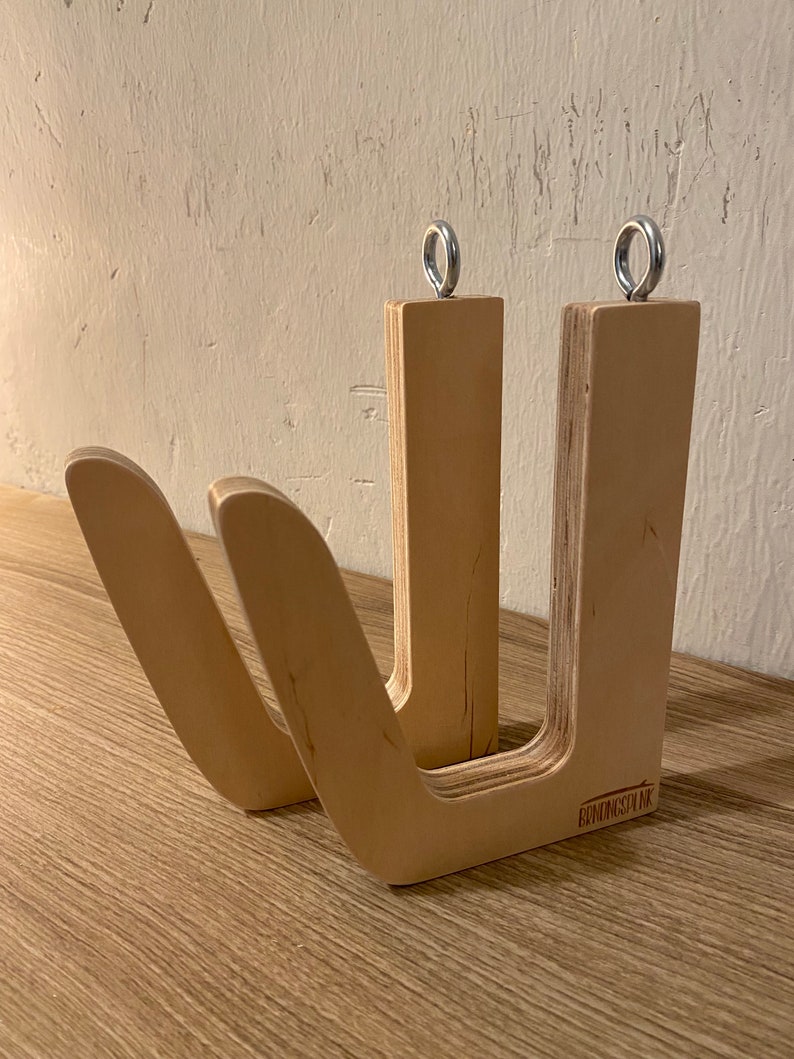 May include: A light-colored wooden surfboard rack with a U-shaped design. The rack has two curved arms and two vertical supports, each topped with a metal ring. The text "BENDINGSPLNK" is visible on the base.