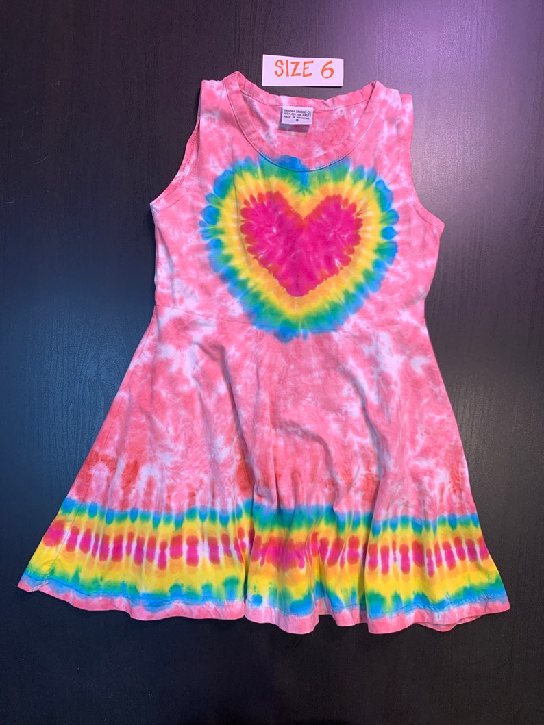 May include: Pink tie-dye sleeveless dress with a rainbow heart design and a yellow, blue, and green tie-dye band around the bottom. The dress has a size tag that says "Size 6".