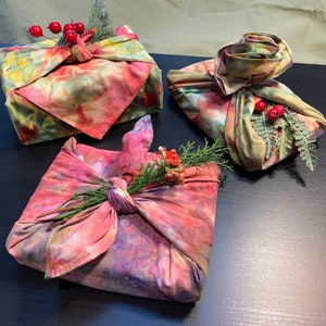 May include: Three fabric-wrapped gifts with a tie-dye pattern. The gifts are wrapped in a traditional Japanese style called Furoshiki. The gifts are decorated with sprigs of greenery and red berries.